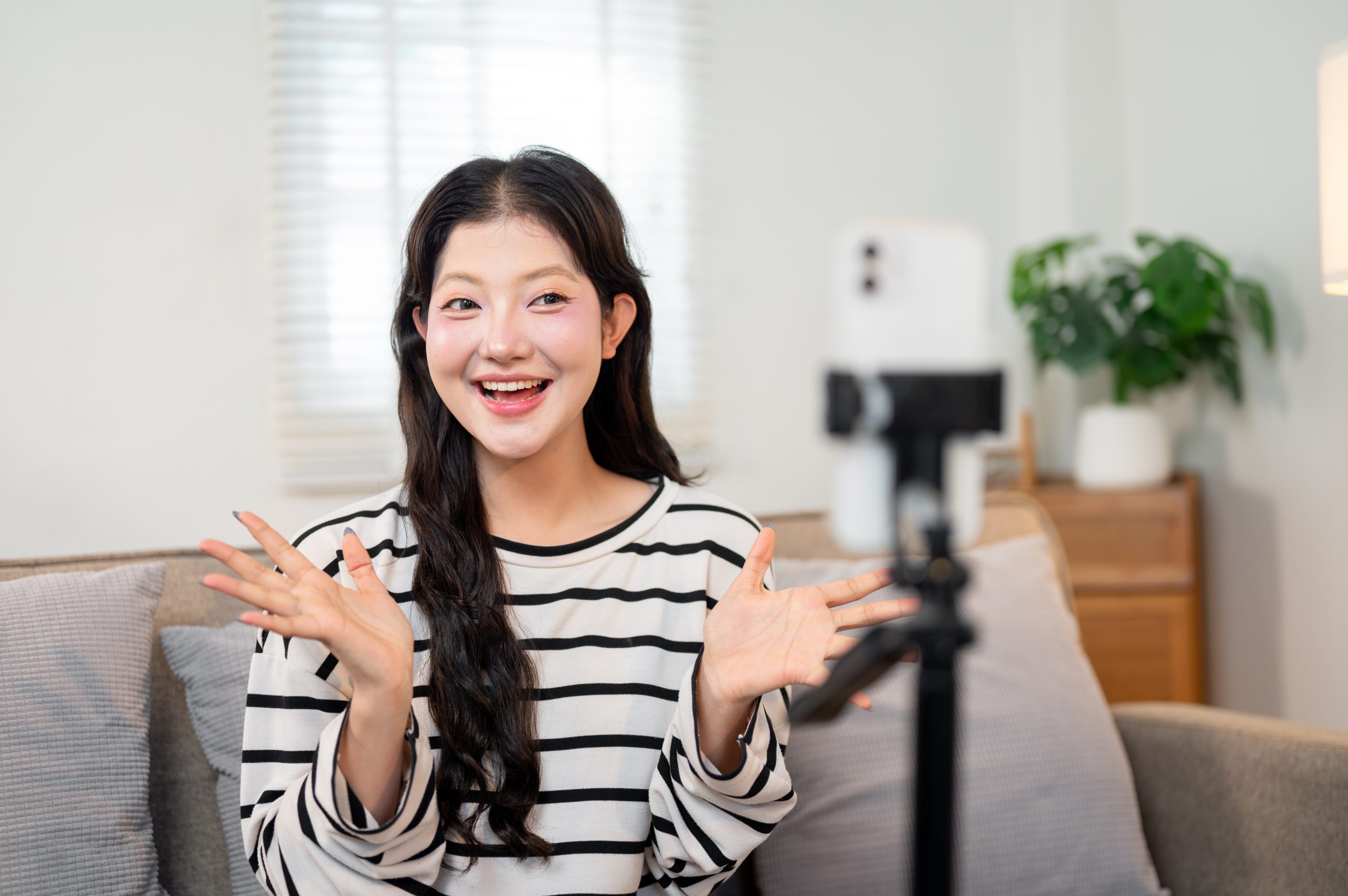 A confident Asian female influencer sits on a sofa, live streaming to engage with her followers.